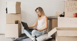 Young woman with laptop sitting surrounded by moving boxes
