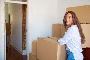 Cheerful beautiful young Hispanic woman unpacking stuff in her new apartment, standing near stacks of carton boxes, looking at camera and smiling.
